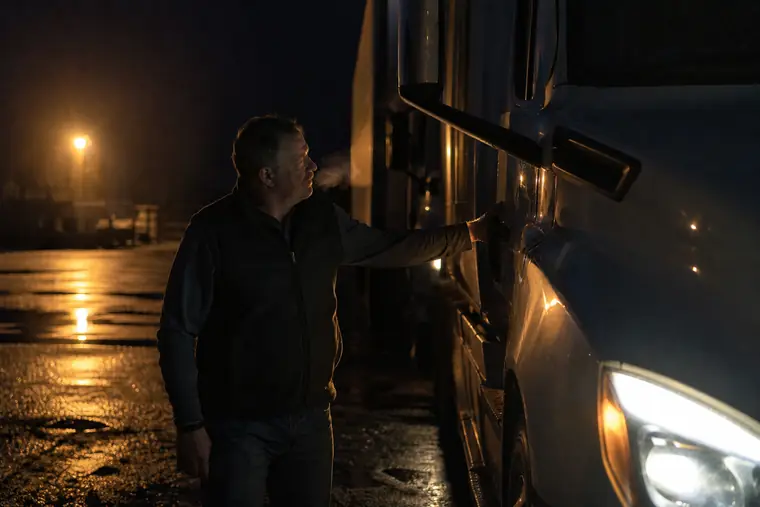 An owner-operator doing a walk-around of a semi truck in a yard before sunrise